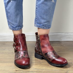 以下が含まれることがあります： フローラルエンボス加工が施された、バーガンディ色のレザーアンクルブーツ。ブーツはサイドジッパーと装飾的なバックルが特徴です。ブーツはローヒールと丸いつま先です。青いジーンズを履いています。