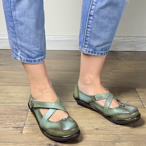 May include: A pair of sage green leather shoes featuring a cross-strap design with embossed floral patterns, a small circular turquoise-colored accent on the toe, and a metallic button detail on the side, paired with blue denim jeans.