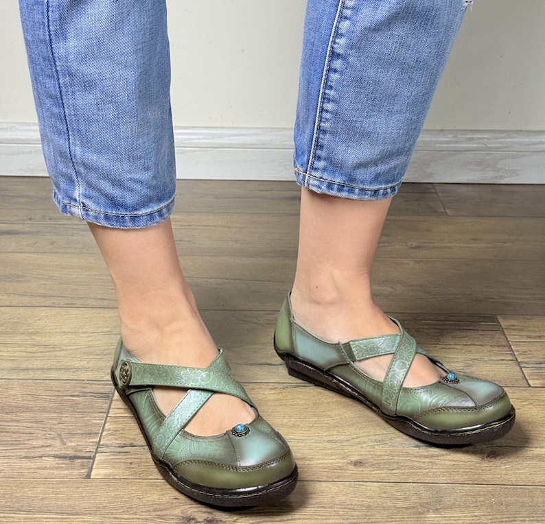May include: A pair of sage green leather shoes featuring a cross-strap design with embossed floral patterns, a small circular turquoise-colored accent on the toe, and a metallic button detail on the side, paired with blue denim jeans.