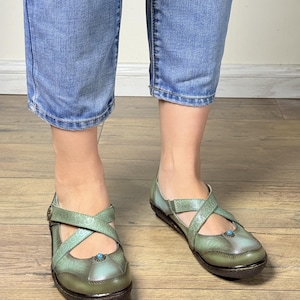 May include: Sage green leather shoes featuring a cross-strap design with embossed floral patterns, a rounded toe, and a small turquoise-colored stone accent at the center. The shoes have a low, dark brown sole and a smooth, polished finish.