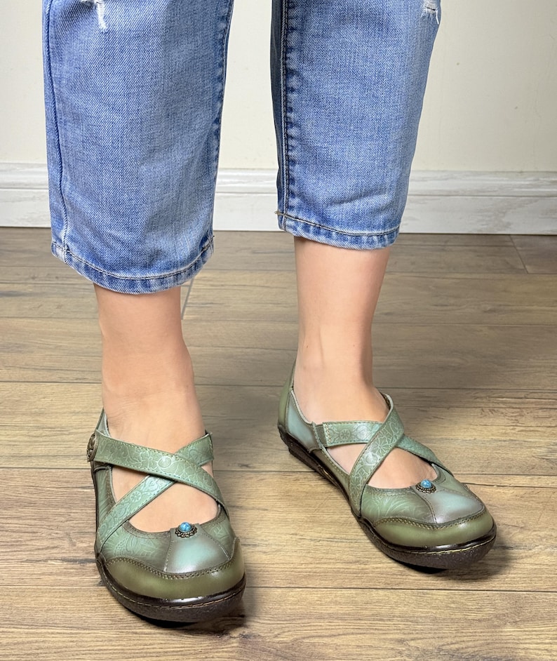 May include: Sage green leather shoes featuring a cross-strap design with embossed floral patterns, a rounded toe, and a small turquoise-colored stone accent at the center. The shoes have a low, dark brown sole and a smooth, polished finish.
