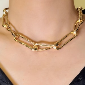 May include: A gold-colored chain necklace with large, textured links. The necklace is worn around the neck and has a chunky, statement design. The links have an irregular shape, giving the necklace a unique, modern look.