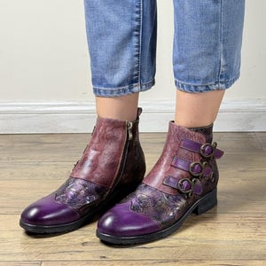 以下が含まれることがあります： 紫とバーガンディ色の革製アンクルブーツ。花と葉の模様が施されています。ブーツにはサイドジッパーと装飾的なバックルが付いています。つま先とかかとは濃い紫色です。