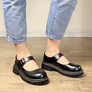 May include: Black Mary Jane shoes with a chunky sole and a buckle strap. The shoes have a glossy finish and a thick, ridged sole. The shoes are worn with light blue denim jeans.