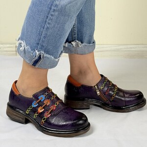May include: Purple leather shoes with colorful stitching and a zipper detail. The shoes have a chunky heel and a rounded toe.