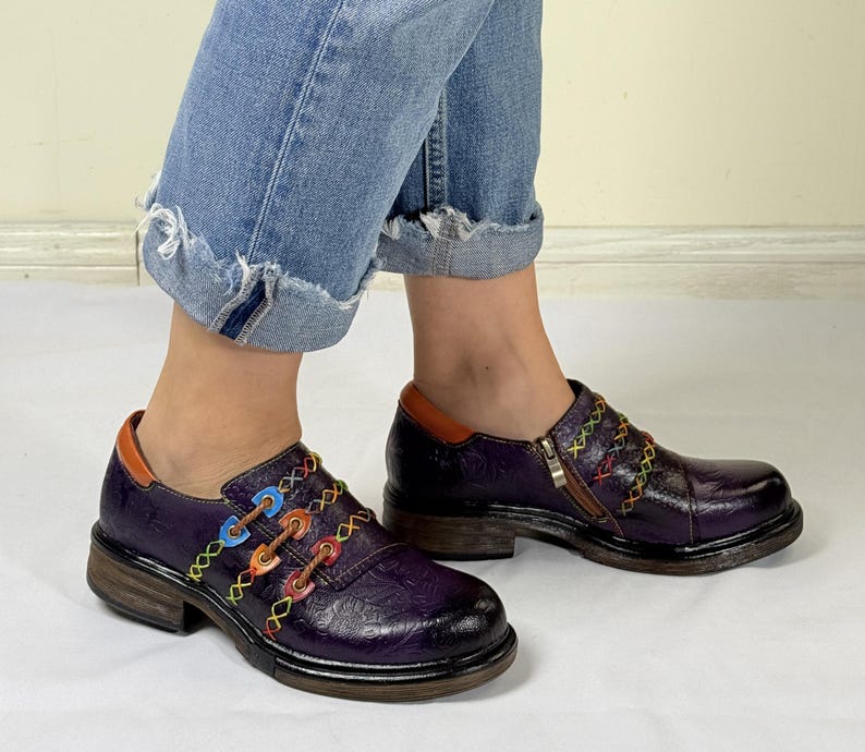 May include: Purple leather shoes with colorful stitching and a zipper detail. The shoes have a chunky heel and a rounded toe.