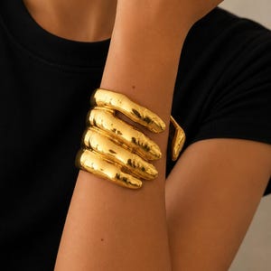 Puede incluir: Un brazalete dorado con forma de mano que se envuelve alrededor de la muñeca. El accesorio metálico presenta cuatro bandas distintas similares a dedos que se curvan alrededor del brazo, creando una apariencia escultural y superpuesta.