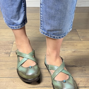 May include: A pair of sage green shoes featuring a cross-strap design across the top, a rounded toe, and a small blue stone accent centered on the vamp. The shoes have a textured, embossed floral pattern and a dark brown sole with a low heel.