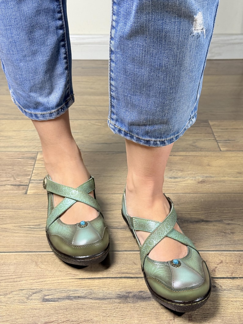 May include: A pair of sage green shoes featuring a cross-strap design across the top, a rounded toe, and a small blue stone accent centered on the vamp. The shoes have a textured, embossed floral pattern and a dark brown sole with a low heel.