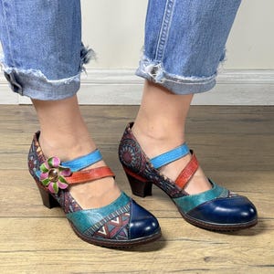 Könnte beinhalten: Bunte Leder-High-Heel-Sandalen mit floralem Akzent. Die Schuhe haben eine blaue Spitze, türkisfarbene und rote Riemen und ein gemustertes Design an den Seiten. Die Absätze sind dunkelbraun, und die Sandalen werden mit einer blauen Jeans getragen.