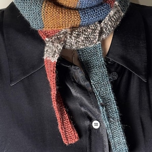 May include: A colorful, knitted scarf with a striped pattern in shades of blue, brown, red, and gray. The scarf is wrapped around the neck and hangs down. The scarf is made of a soft, textured yarn.