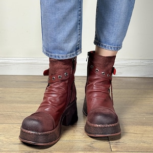 May include: Burgundy ankle boots with a platform sole and a block heel. The boots have a distressed leather appearance, with buckle and stud details. The boots are worn with blue jeans.