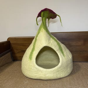 以下が含まれることがあります： ピンクのトップが付いた、薄い緑と白のフェルト製の猫用ベッド。キノコの形をしています。ベッドには猫が出入りできる大きな開口部があります。