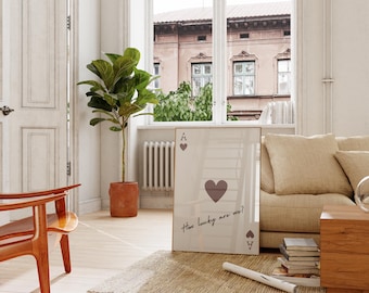 May include: A framed print of a playing card, the Ace of Hearts, leans against a sofa. The card features a large heart symbol and the text "How lucky are we?" The room has a neutral color palette with a wooden chair and a potted plant.