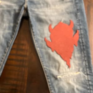 May include: Distressed blue denim jeans with a red devil patch on the right thigh.