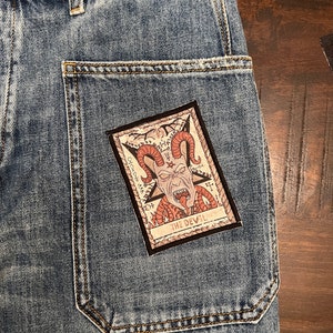 May include: A blue denim jean pocket with a brown and red embroidered patch depicting a tarot card with the text "THE DEVIL" and an illustration of a horned demon with snakes.