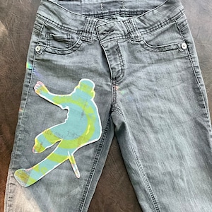 May include: Gray denim jeans with a colorful graphic of a person snowboarding on the left leg.