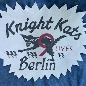 Puede incluir: Parche blanco con una silueta de gato negro y el texto "Knight Kats 9 Lives Berlin".
