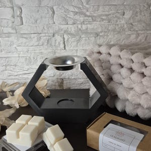 Può includere: Un scaldacera nero geometrico con una ciotola argentata, accanto a cera profumata bianca e una scatola con l'etichetta "TEALIGHT". Lo scaldacera ha un design moderno. Le cere profumate sono in un contenitore di plastica trasparente.
