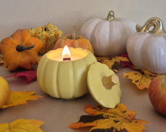 Könnte beinhalten: Eine brennende, gelbe, kürbisförmige Kerze mit einem Holzdocht, umgeben von dekorativen Kürbissen, Herbstblättern und Äpfeln. Zwei weiße Kürbisse mit goldenen Akzenten befinden sich im Hintergrund. Die Szene erinnert an Herbst und Ernte.