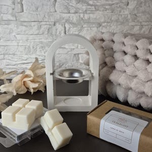 Puede incluir: Un quemador de cera blanco con un plato plateado, acompañado de ceras perfumadas y una caja etiquetada "TEALIGHT CERA DI SOIA". El quemador tiene un diseño arqueado. Las ceras están en un envase de plástico transparente.