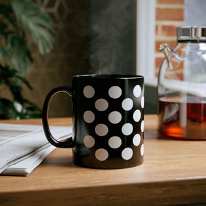 Könnte beinhalten: Eine schwarze Keramiktasse mit weißen Punkten. Die Tasse ist mit einem heißen Getränk gefüllt und Dampf steigt aus der Oberseite auf.