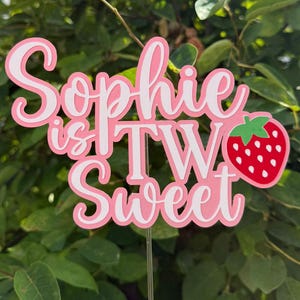 Op de afbeelding: Een taarttopper met de roze en witte tekst "Sophie is TWO Sweet" en een rode aardbei-afbeelding met witte zaadjes. De topper is bevestigd aan een dun, doorzichtig, verticaal stokje om in een taart of ander voedsel te steken.