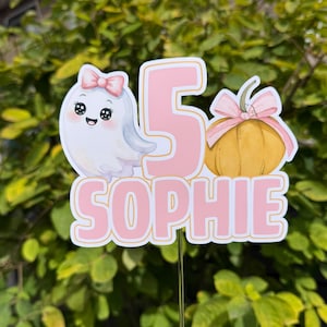 Op de afbeelding: Een taarttopper met een cartoonspookje met een roze strik, een groot cijfer 5 en een gele pompoen met een roze strik. De naam SOPHIE staat onderaan in grote roze letters gedrukt. Het ontwerp is bevestigd op een doorzichtig, dun plastic stokje.
