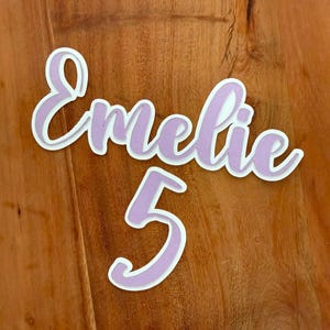 Op de afbeelding: Een gepersonaliseerd uitgesneden bord met de naam Emelie in een cursief lettertype, geplaatst boven het cijfer 5. De letters en het cijfer zijn uitgevoerd in een lichtpaarse kleur met een dikke witte rand voor een gelaagd effect.