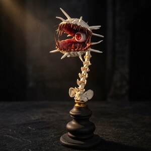 Könnte beinhalten: Eine Skulptur mit einem weißen, stacheligen, muschelartigen Kopf, einem einzelnen roten Auge und scharfen Zähnen, montiert auf einer weißen Wirbelsäule. Das gesamte Objekt steht auf einem schwarzen, gestuften Sockel mit goldenem Ring.