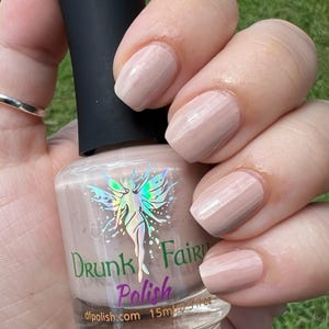 May include: A bottle of light pink nail polish with a holographic fairy design and the text "Drunk Fairy Polish" on the label. The bottle is 15 ml or 0.5 fl oz. The polish is shown on a hand with five nails painted in a light pink color.