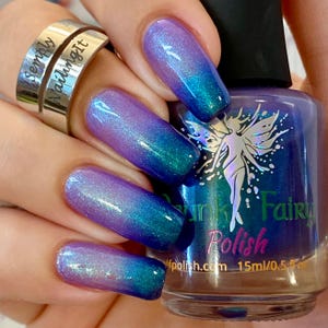 May include: A hand displays nails painted with a gradient of purple, teal, and blue nail polish. A bottle of "Funk Fairy Polish" is visible, featuring a fairy illustration. A silver ring with the words "Simply Nailing It" is also in the image.