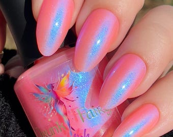 May include: Close-up of long, oval-shaped nails painted with a pink iridescent nail polish with a blue shimmer. The nail polish bottle is pink with a fairy graphic and the words "Drunk Fairy Polish".