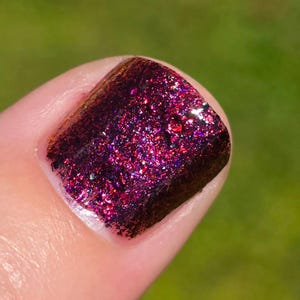 May include: Close-up of a fingernail painted with a dark burgundy nail polish. The polish is heavily textured with sparkling glitter in shades of red, pink, and purple. The background is a blurred green.