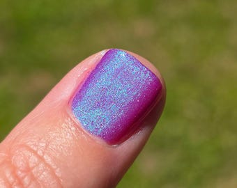 May include: Close-up of a fingernail painted with iridescent nail polish. The polish shifts between shades of purple and blue, with a shimmering, glittery effect. The nail is well-manicured and reflects sunlight.