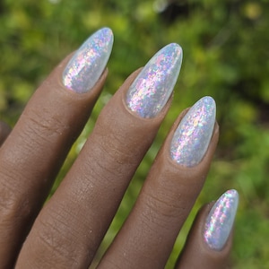 May include: Close-up of a hand with long, oval-shaped nails painted with iridescent, shimmering nail polish. The polish has a light, pearlescent base with colorful, glittery flecks. The background is a blurred green.