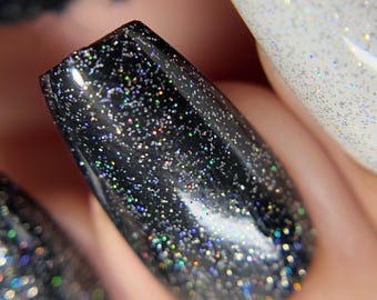 May include: Close-up of two fingernails painted with black and white glitter nail polish. The black glitter nail polish has a holographic finish.