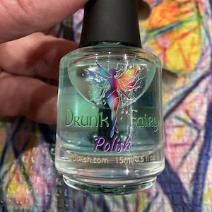 May include: A clear glass bottle of nail polish with a green iridescent shimmer. The bottle has a black cap and a label that reads "Drunk Fairy Polish" with a fairy illustration. The label also includes the website "dfpolish.com" and the size "15ml/0.5 fl oz."