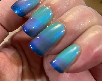 May include: Close-up of a hand with manicured nails. The nails are painted with a gradient effect, transitioning from a deep blue at the tips to a teal green towards the base. The polish has a glossy, iridescent finish.
