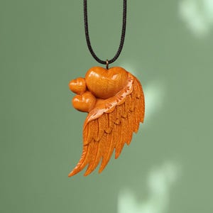 May include: A wooden pendant necklace featuring a carved heart and wing design. The pendant is a light orange colour and hangs from a black cord.