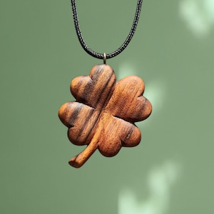 May include: A wooden four-leaf clover pendant on a dark cord necklace. The clover is carved from light brown wood with darker grain patterns. The pendant hangs against a soft green backdrop.