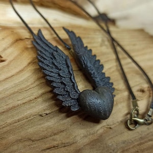 May include: A black wooden heart pendant with wings, hanging from a black cord necklace. The pendant is carved with intricate details and has a rustic, gothic aesthetic.