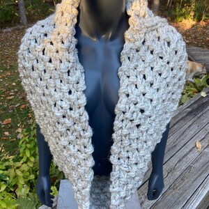 May include: A cream-coloured, hand-knitted cowl scarf. The scarf is made of thick yarn with a textured, open weave design. The cowl is displayed on a mannequin, showcasing its shape and drape.