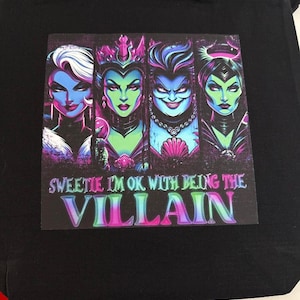May include: Black tote bag featuring a colorful graphic of four stylized female villains with the text "Sweetie I'm Ok With Being The Villain". The graphic has a purple, green, and blue color scheme.