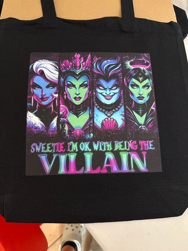 May include: Black tote bag featuring a colorful graphic of four stylized female villains with the text "Sweetie I'm Ok With Being The Villain". The graphic has a purple, green, and blue color scheme.