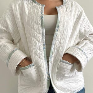 May include: A white quilted jacket with a floral patterned trim and pockets. The jacket has a quilted design and a round neckline. The trim features a light blue and green floral pattern. The jacket is worn open.