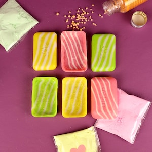 Puede incluir: Seis jabones coloridos con diseños en espiral en amarillo, rosa y verde, dispuestos sobre una superficie púrpura. También hay pequeños paquetes de polvo de color y una botella de gránulos, lo que sugiere un proyecto de manualidades o bricolaje.