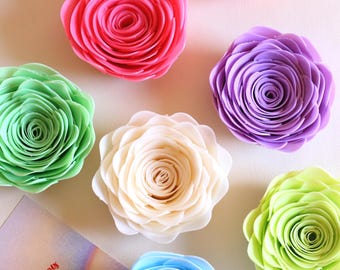 May include: A selection of artificial rose flowers in various colours, including pink, orange, purple, green, white, and blue. The roses are arranged on a white surface. A card with the text "MAY THIS DAY BE SPECIAL!" is also visible.