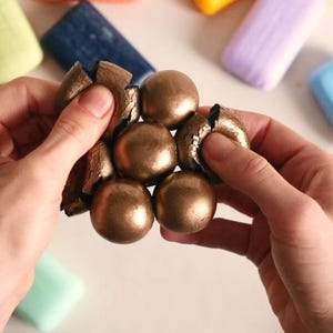 May include: Hands holding a cluster of six metallic gold spheres, some cracked open to reveal a textured interior. Various colourful soap bars in orange, yellow, blue, and purple are scattered in the background on a white surface.