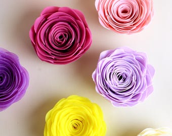 May include: A selection of artificial rose flowers in various colours. The roses are in shades of red, pink, purple, yellow, and cream. The petals are detailed and layered, giving a realistic look. The flowers are arranged on a light background.